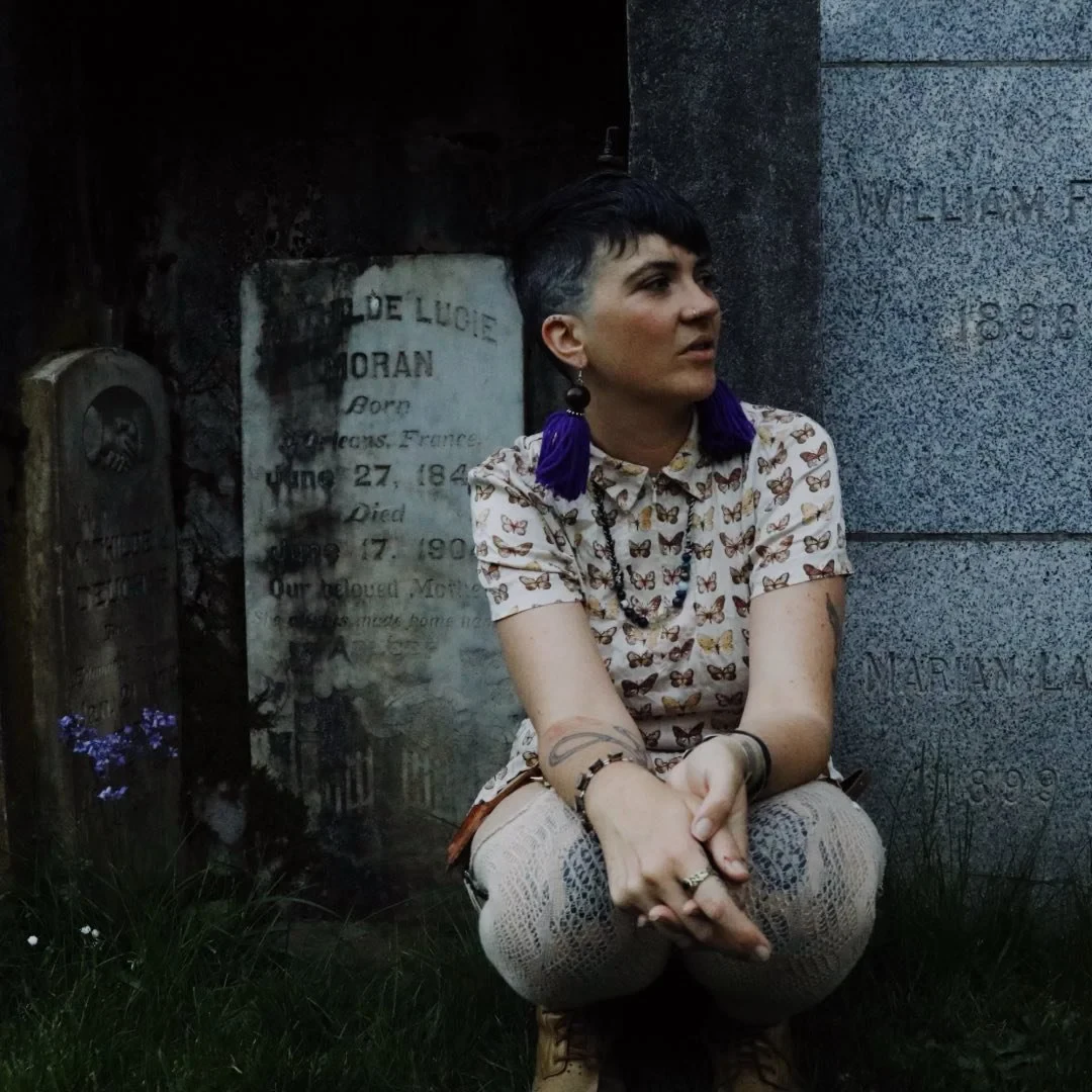 Olivia Pepper sitting on the grass in a cemetery between weathered headstones, wearing a butterfly-print shirt and long purple tassel earrings, looking off to one side.