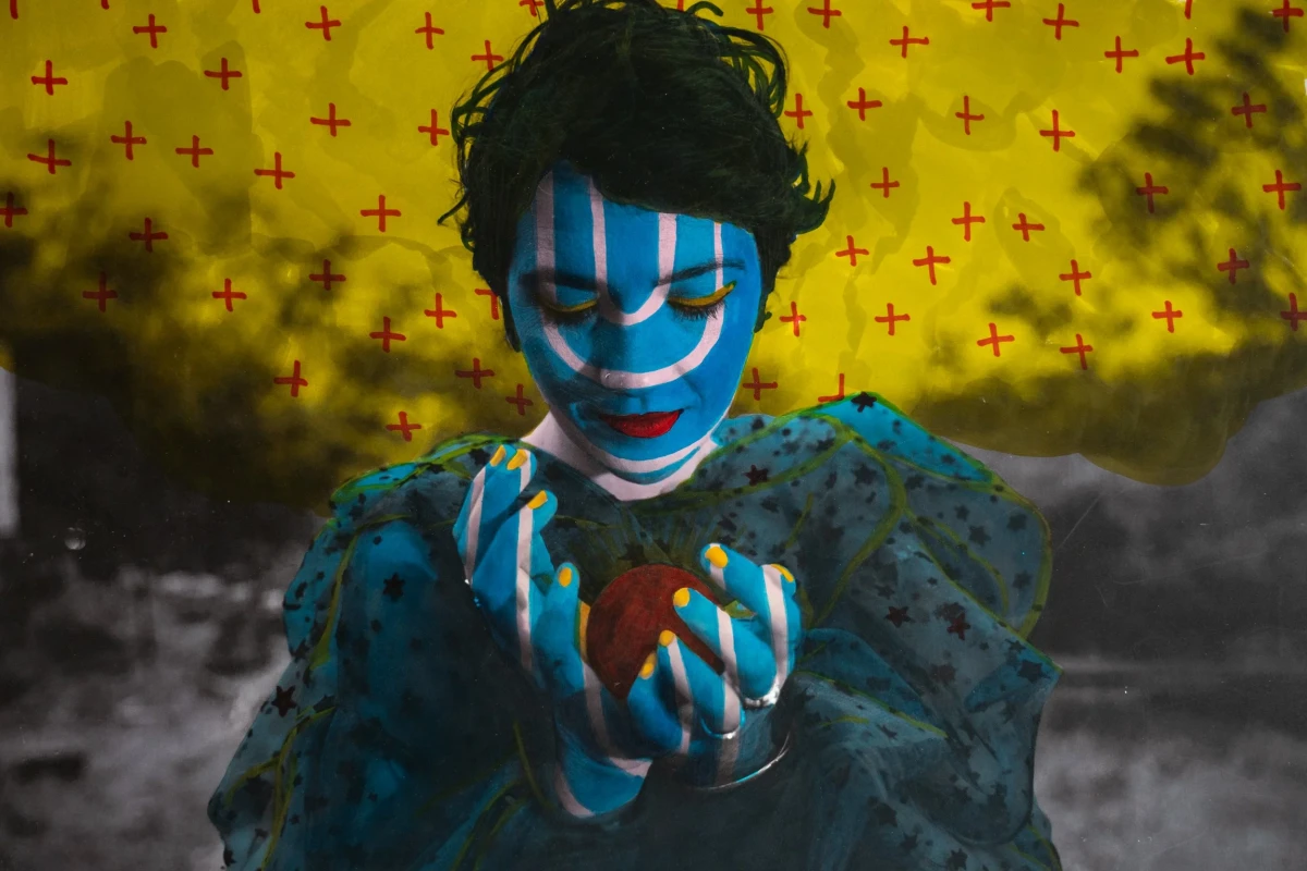 Hand-coloured photograph of Olivia Pepper with face and hands painted blue and white, cradling a red apple, against a yellow sky stencilled with small red crosses.