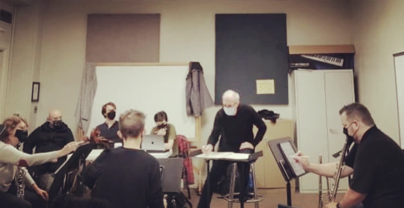 Candid rehearsal photo: masked players — clarinet, flute, cello and others — seated in a rehearsal room around a conductor perched on a stool, score open on a stand in front of him.