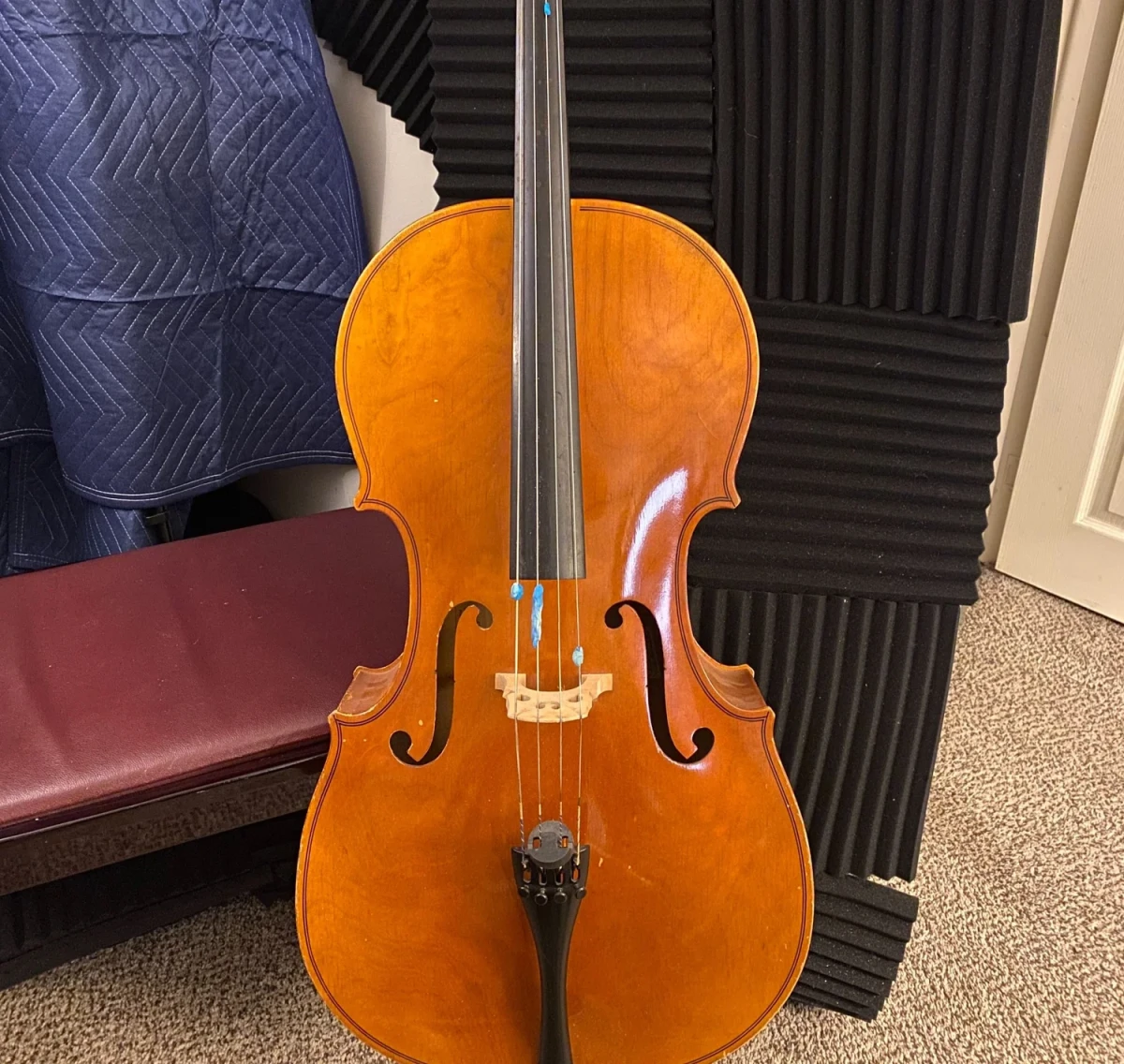 A cello standing upright against acoustic foam, with lumps of blue Blu-Tack pressed onto the strings between the bridge and the end of the fingerboard.